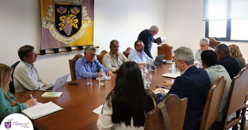 A group of professionals seated around a long conference table in a meeting room, some taking notes and using laptops, with a banner featuring a coat of arms on the wall behind them. Carregal do Sal investe 1,8 milhões de euros em novo parque de lazer no centro da vila