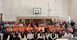 Group portrait in a gym: seniors in orange shirts seated in front with student musicians in formal attire kneeling at the front; banner reads Politécnico de Viseu on the right.]','Large crowd of seniors in orange shirts posing with younger musicians in front of a school gym; basketball hoop visible in the background; banner Politécnico de Viseu on the right.','Multi-generational group photo at a school event in a gym, featuring elderly volunteers in orange shirts and a row of young musicians in white shirts and black vests in front.','Group photo inside a gymnasium showing an audience in bright orange shirts and a band of young musicians in front; a banner for Politécnico de Viseu is visible.','Crowd shot from a school event: elderly participants in orange shirts with a performance group of students in front, banner reading Politécnico de Viseu on the side.