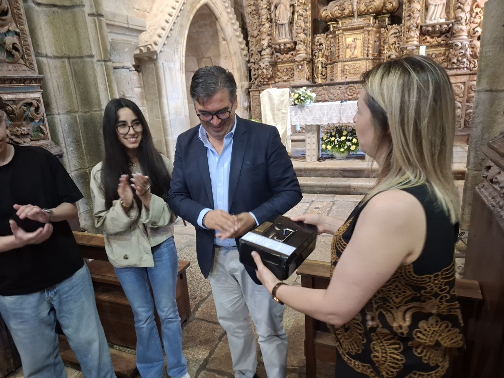 Man in a blue blazer receiving a boxed item from a smiling presenter inside a ornate stone church, while two young people clap in the background. Mosteiro de Cárquere recebe “Caixa do Tempo” para ser aberta em 2046