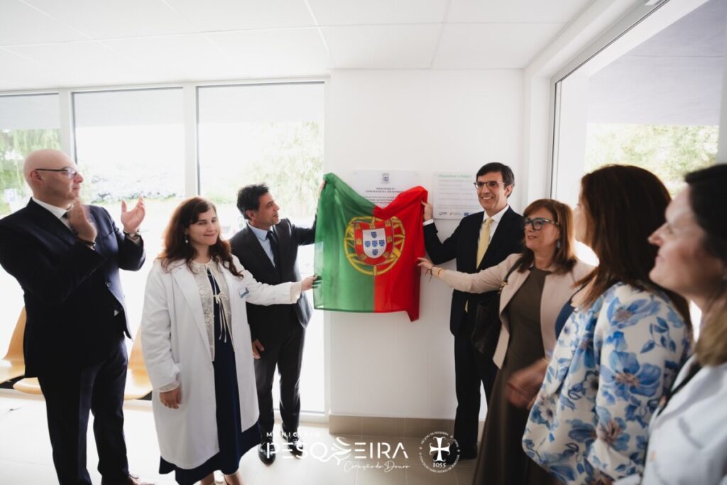 Group of professionals in formal attire unveiling a Portugal flag on a white wall during a ceremony, smiling and clapping in a bright office space. Câmara de São João da Pesqueira quer reforço médico e Serviço de Urgência Básica