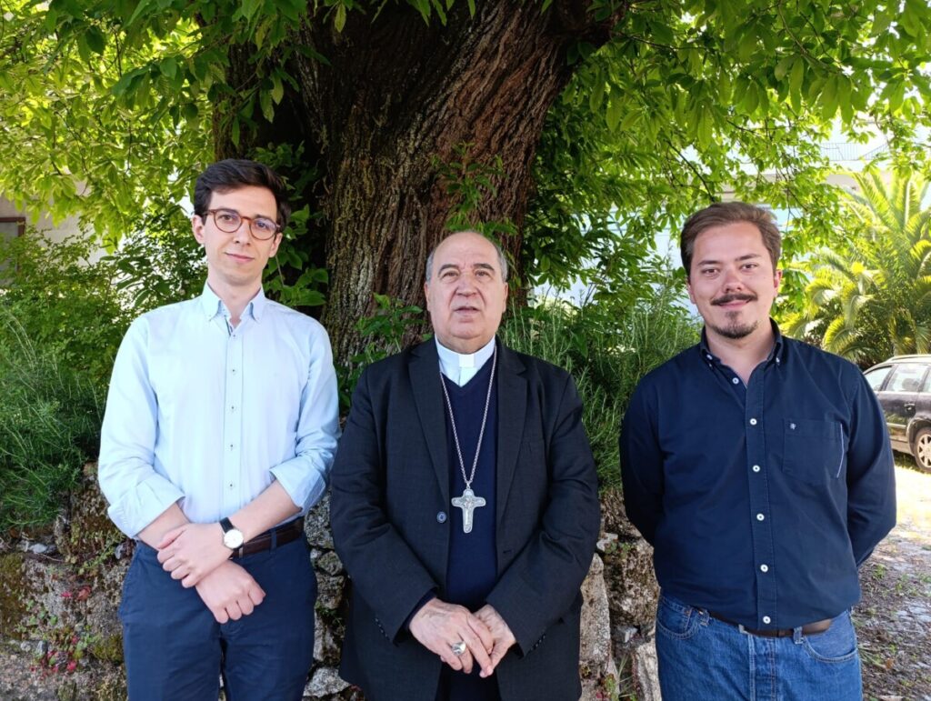 Three men pose outdoors in front of a large tree; the center man is a priest wearing a clerical collar and a cross necklace. Tiago Rio e Francisco Ferreira ordenados diáconos no próximo domingo na Diocese de Viseu