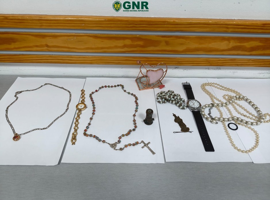 Assorted jewelry including necklaces, a watch, rosary, and trinkets laid out on white sheets for display, with a GNR logo in the background. Ladrão apanhado em flagrante após onda de furtos em residências de Viseu e de Penalva do Castelo