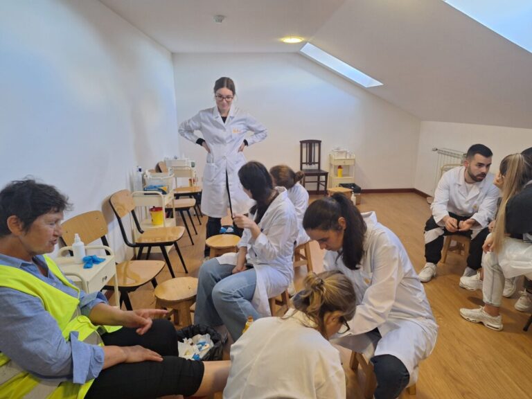 Medical training session: students in white coats treat a seated man wearing a yellow safety vest while a supervisor stands nearby in a white coat.