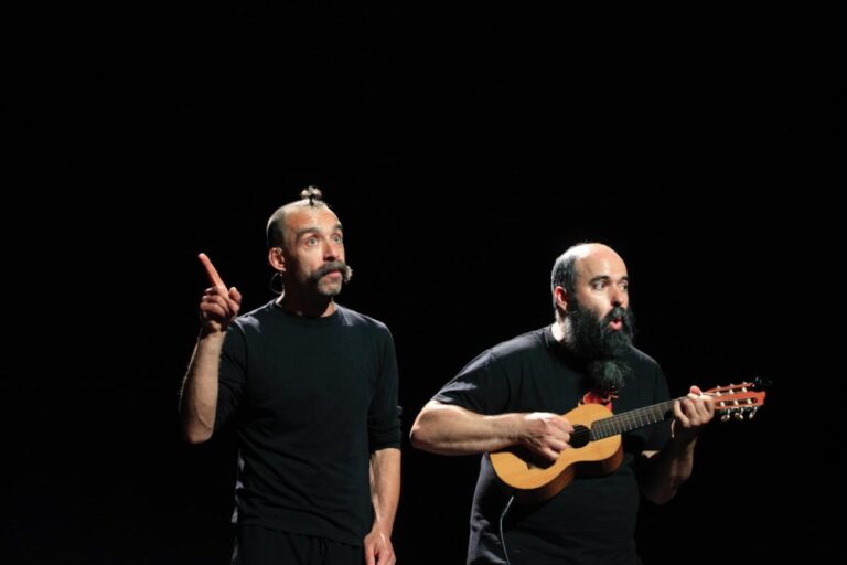 Two male performers on a dark stage: one points upward while the other plays a small guitar (ukulele).