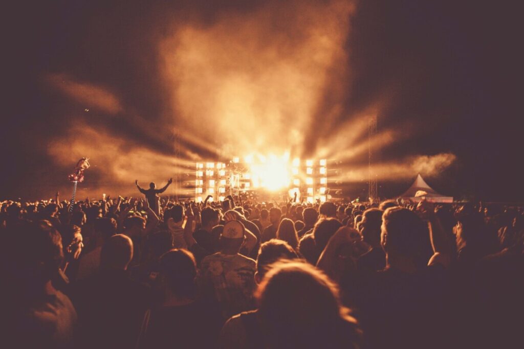 Crowd enjoying an outdoor concert at night with a bright, illuminated stage and smoky beams in the air Viseu vai ter festival “Rock in Dão” em setembro