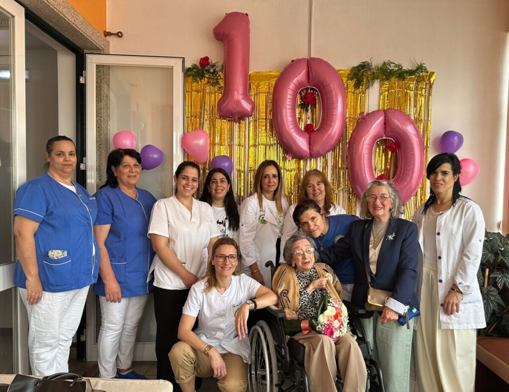 Group of nurses and caregivers pose with an elderly woman in a wheelchair in front of pink '100' balloons and gold streamer backdrop, celebrating a milestone. Utente do Centro Social Paroquial de Rio de Loba celebra 100 anos de vida