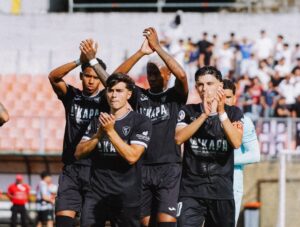 Group of soccer players in black uniforms standing together on the field, clapping after a game, with blurred stands in the background.