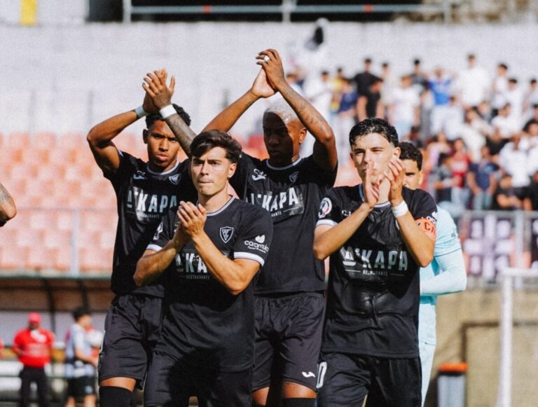Group of soccer players in black uniforms standing together on the field, clapping after a game, with blurred stands in the background.