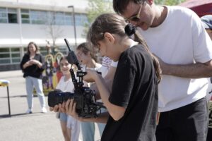 Girl in black guides a professional camera on a stabilizer at an outdoor school event, with a man nearby.