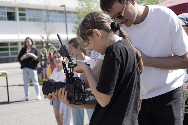 Girl in black guides a professional camera on a stabilizer at an outdoor school event, with a man nearby.