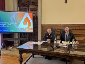 Two men sit at a polished wooden conference table in a historic room; one in a suit and the other in a uniform, with a large colorful presentation screen to the left.