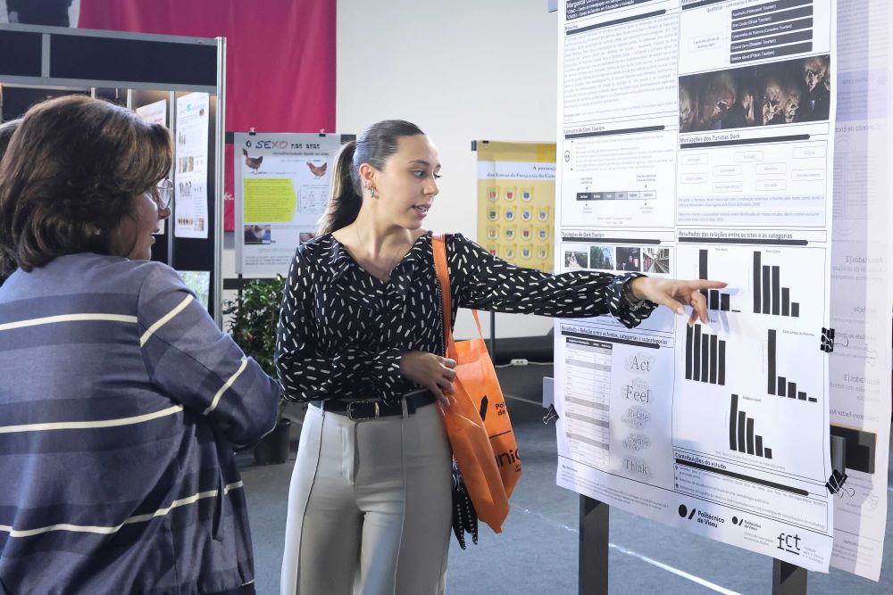 Woman presenting a scientific poster to another attendee at a conference, pointing to bar charts and graphs on the board with an orange tote bag worn over her shoulder by the presenter in a black patterned blouse and light pants. Instituto Politécnico de Viseu abre em maio as portas à comunidade