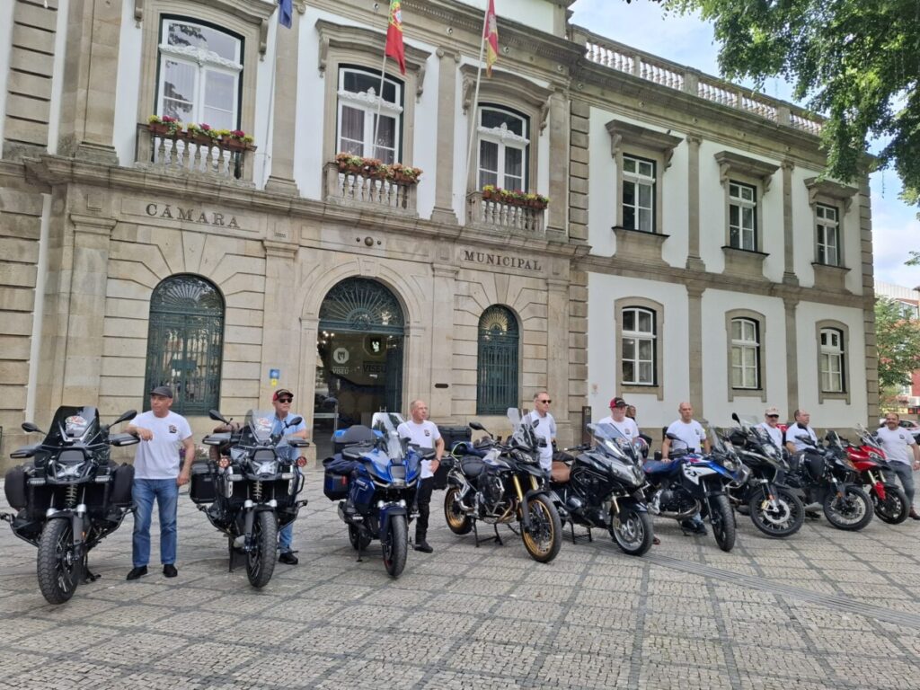 Line of motorcycles parked in front of a historic municipal building, with riders in white shirts standing beside them on a cobblestone plaza. Grupo de 10 motociclistas de Viseu parte rumo a Marrocos numa aventura de 17 dias