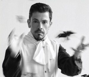 Black-and-white photo of a male magician in a white cravat, tossing playing cards mid-gesture with a focused expression.