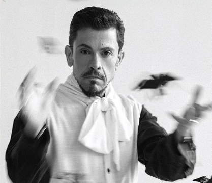 Black-and-white photo of a male magician in a white cravat, tossing playing cards mid-gesture with a focused expression.