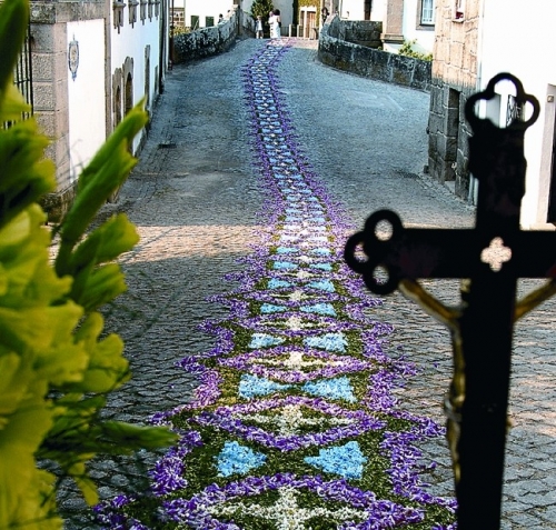 Cobblestone street adorned with a purple and white flower carpet; a crucifix in the foreground hints at a religious procession.
