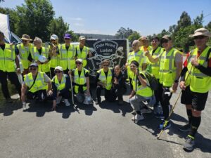 Group of people in bright safety vests posing outdoors for a group photo with a biker club banner in the background, smiling and giving thumbs up.