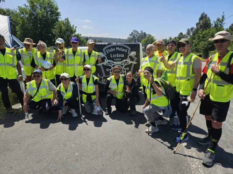Group of people in bright safety vests posing outdoors for a group photo with a biker club banner in the background, smiling and giving thumbs up.