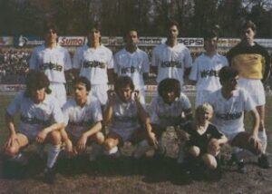 Soccer team posing for a pre-match group photo on a field, players in white uniforms with dark socks, plus a child in front, with banners in the background.