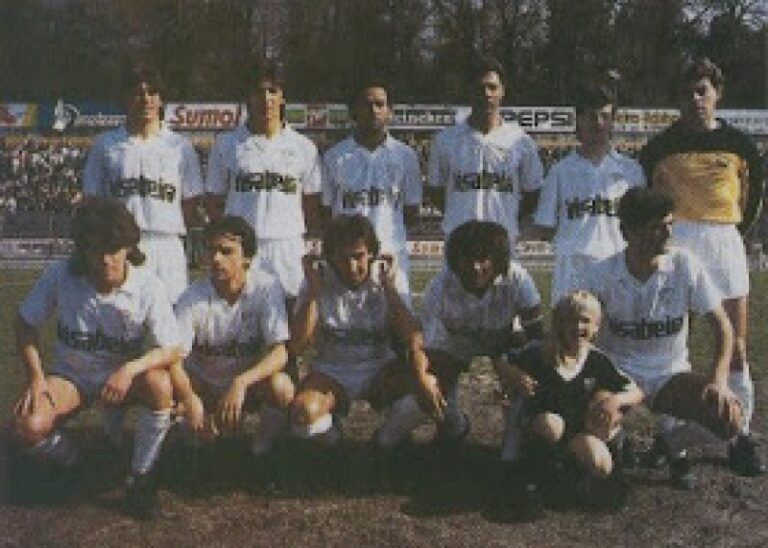 Soccer team posing for a pre-match group photo on a field, players in white uniforms with dark socks, plus a child in front, with banners in the background.