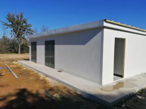 Small modern prefab building under construction with white corrugated metal siding, glass doors, and a concrete walkway on a dirt lot under a blue sky