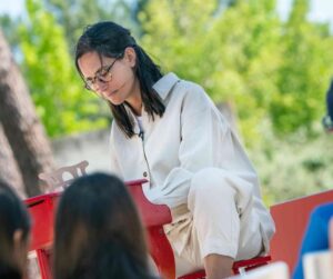 Woman in white outfit and glasses playing a red outdoor piano, with green trees in the background.