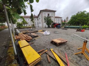 Construction site in a courtyard with piles of wood, yellow boards, pallets, and gravel, in front of a white multi-story building under overcast skies.
