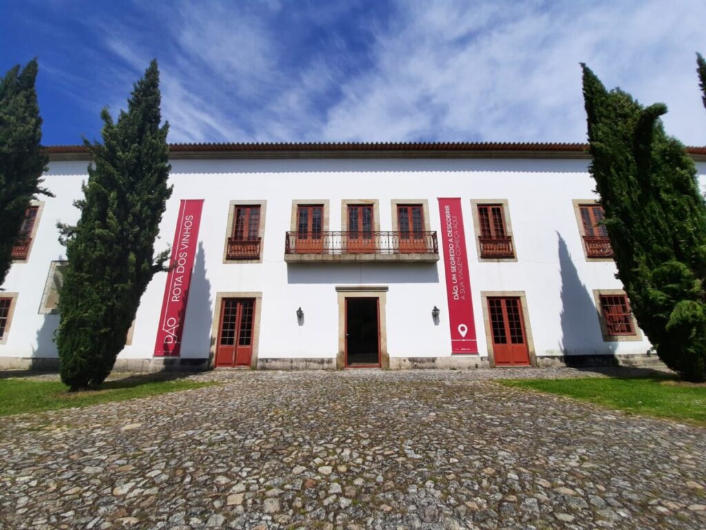 White two-story building with red-trimmed windows and doors, red banners on either side, and a cobblestone foreground flanked by tall evergreen trees under a blue sky. Solar do Dão recebe nova edição das “Tertúlias do Fontelo” com António Martins da Cruz
