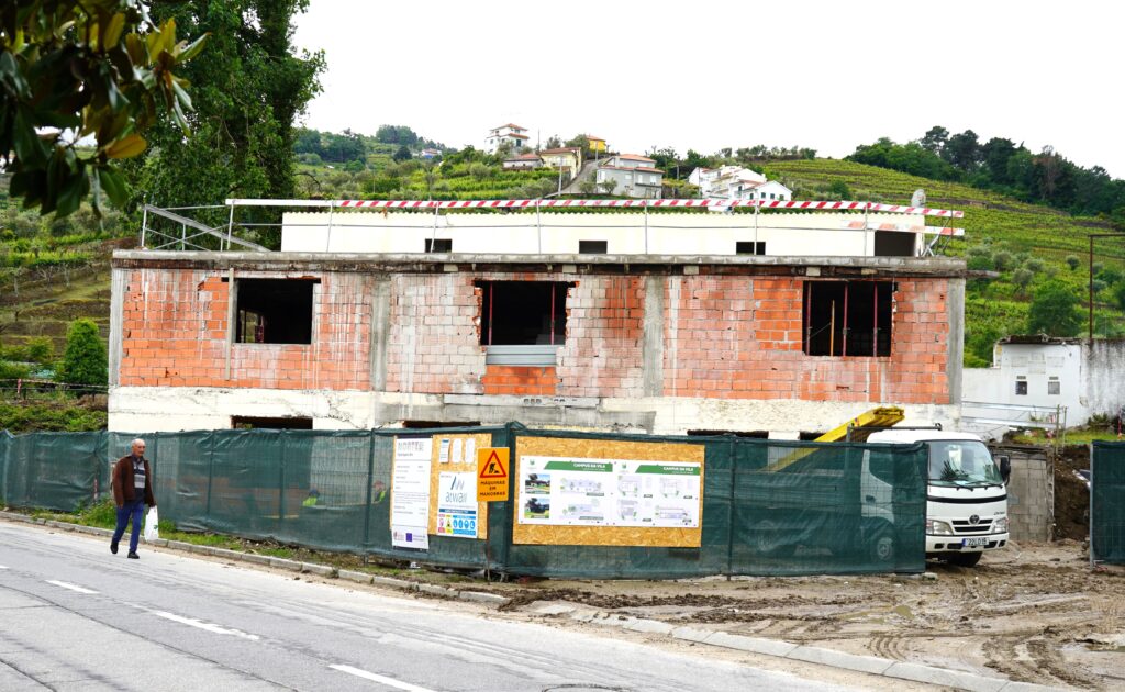 Unfinished brick building under construction behind a green construction fence, with a man walking on the street in the foreground. Lamego reabilita edifício degradado em Cambres para criar centro cívico