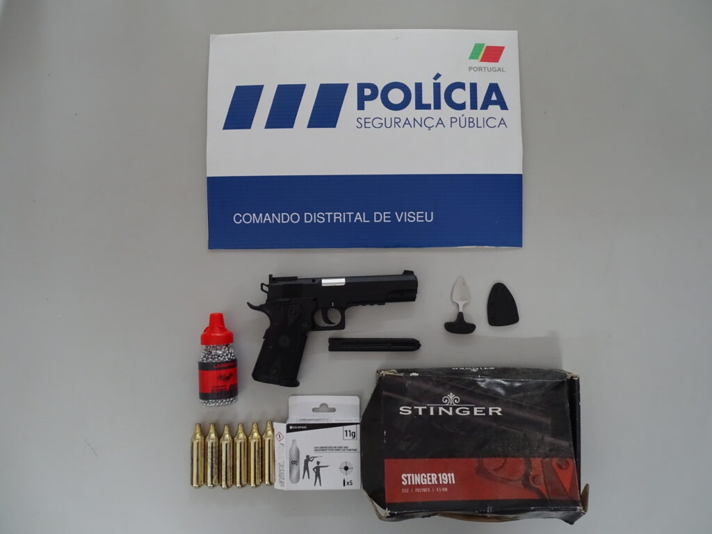 Police sign on wall reading 'POLICIA SEGURANÇA PÚBLICA' with blue lower panel saying 'COMANDO DISTRITAL DE VISEU'; handgun and ammunition laid out on a table shelf below. PSP apreende arma de ar comprimido e arma proibida em operação em Viseu