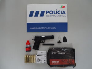 Police sign on wall reading 'POLICIA SEGURANÇA PÚBLICA' with blue lower panel saying 'COMANDO DISTRITAL DE VISEU'; handgun and ammunition laid out on a table shelf below.