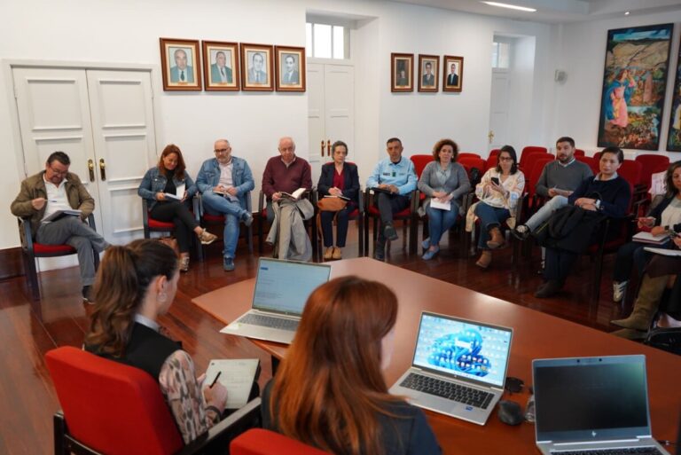 Group of diverse adults seated in a circle in a bright meeting room, with laptops on a central table foregrounded for discussion accessions.