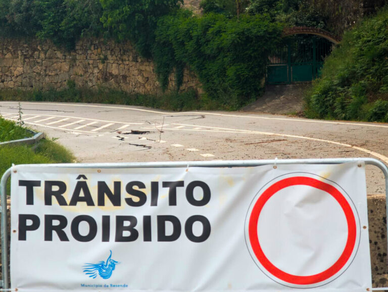 Road closed barrier with a banner reading 'TRÂNSITO PROIBIDO' and a large red no-entry circle, with a gated, tree-filled backdrop.