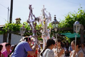 Outdoor religious procession with people carrying ornate crosses and lanterns adorned with flowers under a sunny sky.
