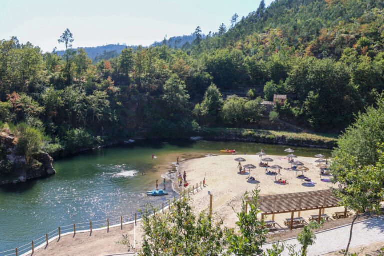 A sunny riverside beach with people sunbathing under straw umbrellas on a sandy shore, next to a calm green river framed by forested hills.