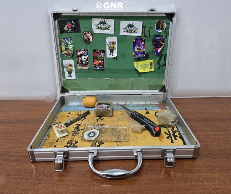 Open metal briefcase on a wooden table containing small items: a black-handled knife, rocks, a small bag, and scattered paraphernalia; stickers cover the green interior lid labeled GNR in the background.