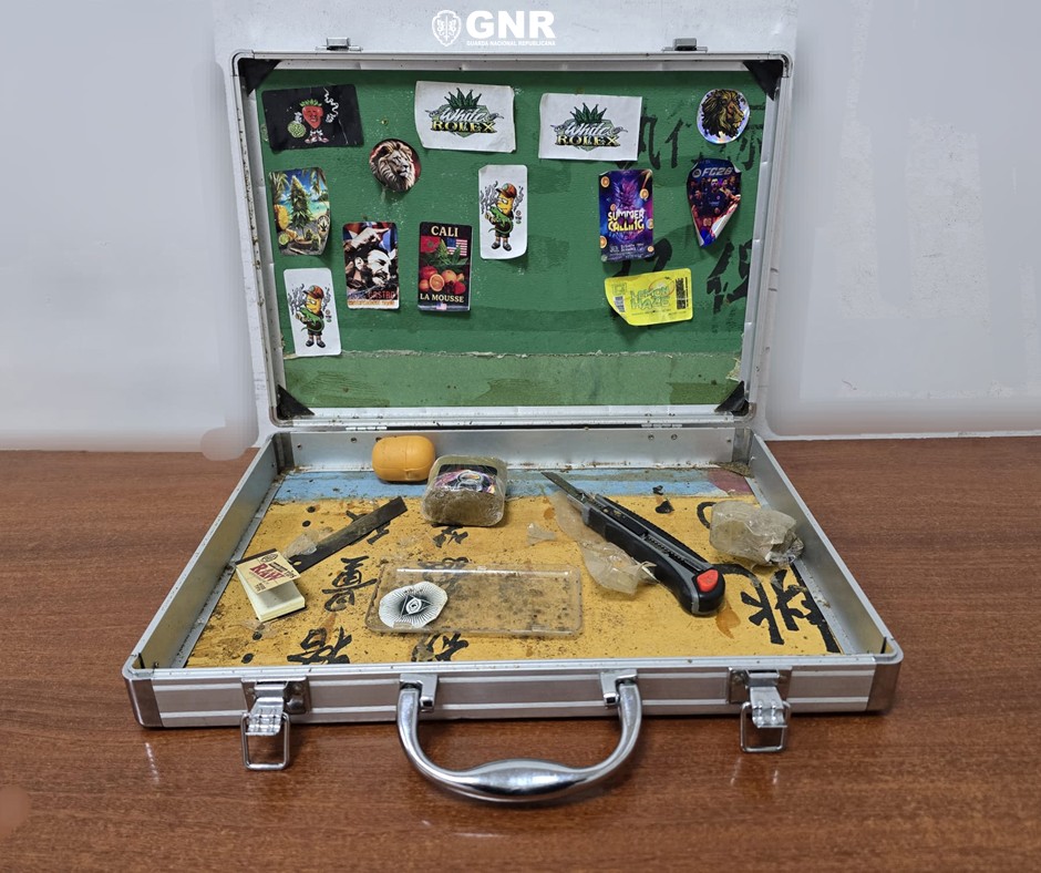 Open metal briefcase on a wooden table containing small items: a black-handled knife, rocks, a small bag, and scattered paraphernalia; stickers cover the green interior lid labeled GNR in the background. Homem detido por tráfico de droga em operação da GNR em Viseu
