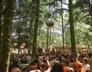 Crowd dancing at an outdoor forest party with a DJ booth and hanging lanterns among trees.