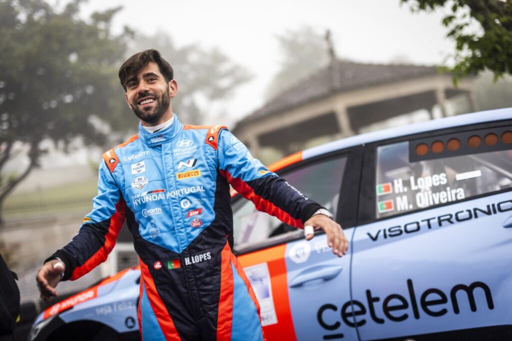 Smiling race car driver in a blue and orange suit stands beside a blue race car with sponsor decals. Hugo Lopes aposta forte no Rally de Lisboa para relançar temporada