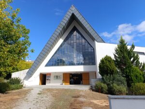 Modern church with a tall triangular glass facade and open wooden doors under a sharp pitched roof, blue sky above.