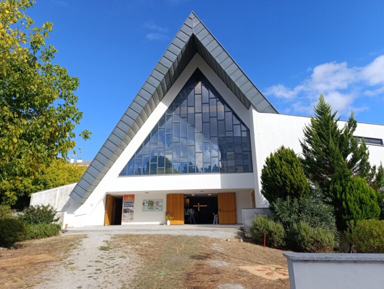 Modern church with a tall triangular glass facade and open wooden doors under a sharp pitched roof, blue sky above.