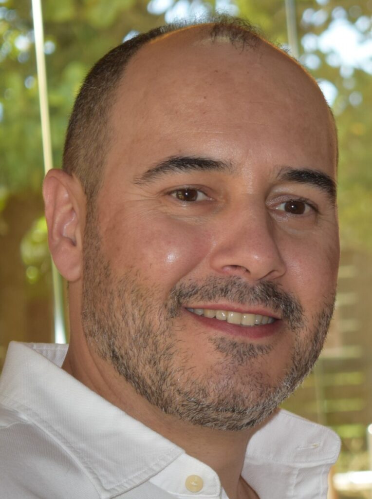 Close-up portrait of a smiling man with short hair and light stubble, wearing a white button-up shirt, indoors by a window with greenery behind. Parentalidade em mudança
