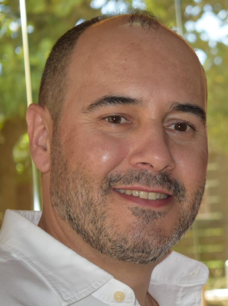Close-up portrait of a smiling man with short hair and light stubble, wearing a white button-up shirt, indoors by a window with greenery behind.