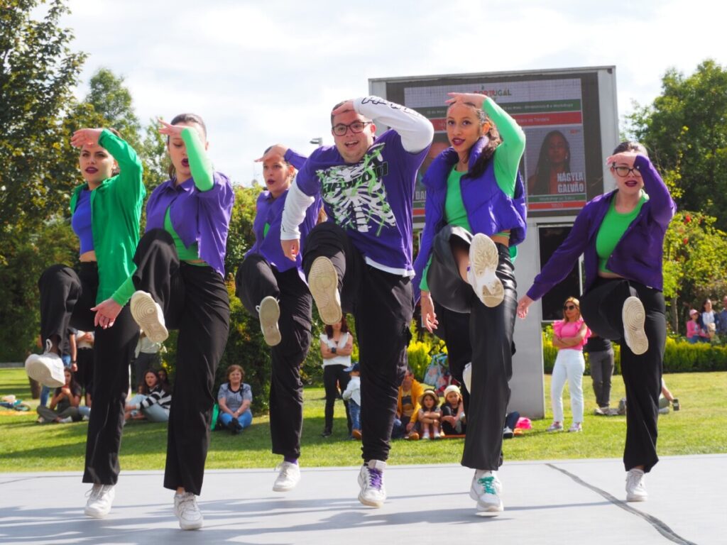 Group of dancers performing a synchronized kick on an outdoor stage, wearing green tops and purple jackets with white sneakers, audience watching in the background. Tondela recebe primeira eliminatória do Portugal a Dançar com 176 bailarinos