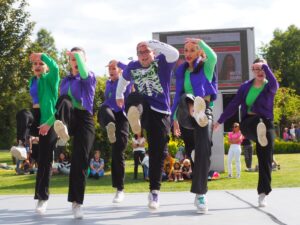 Group of dancers performing a synchronized kick on an outdoor stage, wearing green tops and purple jackets with white sneakers, audience watching in the background.