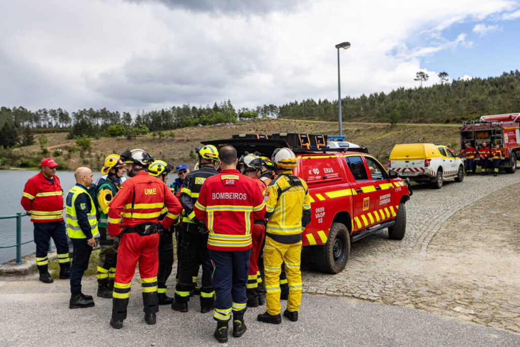 Group of firefighters and emergency responders in red and yellow gear chat near a red rescue SUV by a cobbled road beside a lake and hillside.