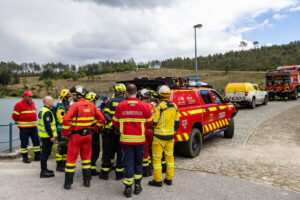 Group of firefighters and emergency responders in red and yellow gear chat near a red rescue SUV by a cobbled road beside a lake and hillside.