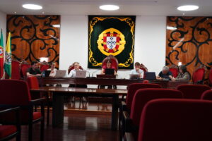 Panel of officials seated at a long desk in a formal meeting room, Portuguese flag on the left and a large crest banner behind them.