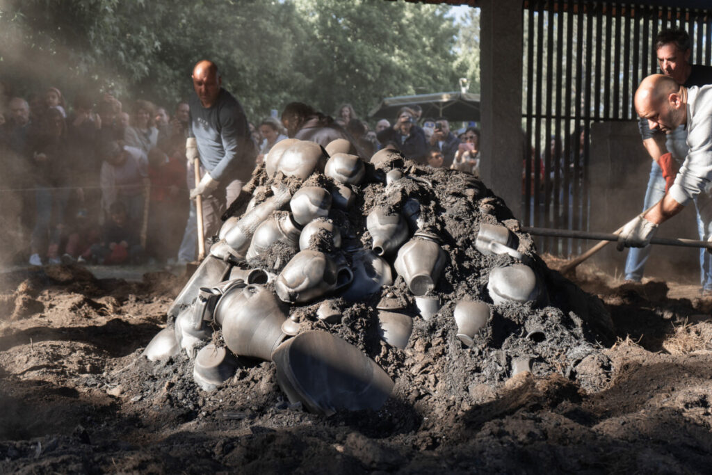 Pile of blackened metal pots and ash being dug from a mound while a crowd watches a public demonstration.