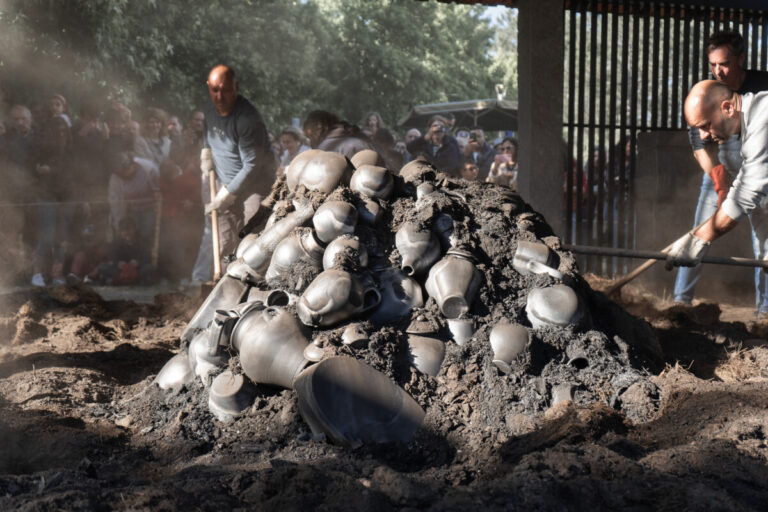 Pile of blackened metal pots and ash being dug from a mound while a crowd watches a public demonstration.